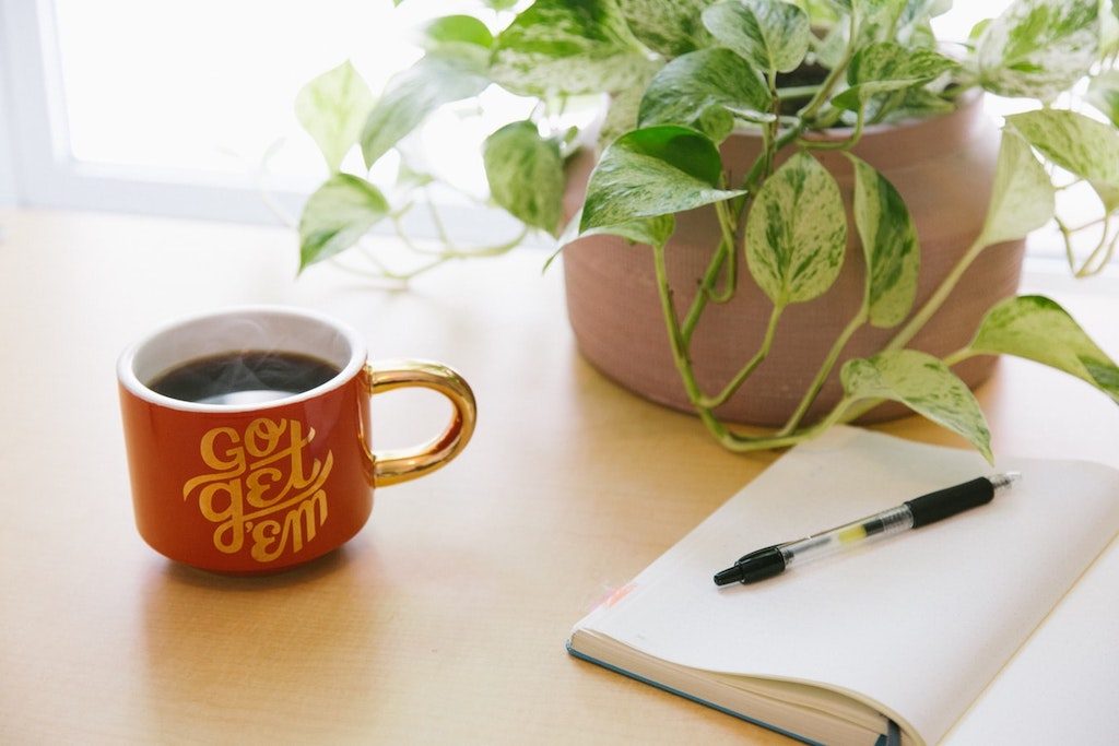 coffee cup that says "go get em" next to notebook with pen, plant